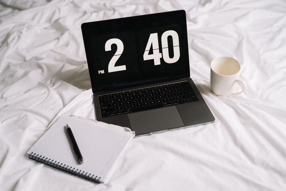 Laptop displaying time next to a notebook and coffee on a bed, creating a cozy work setup.